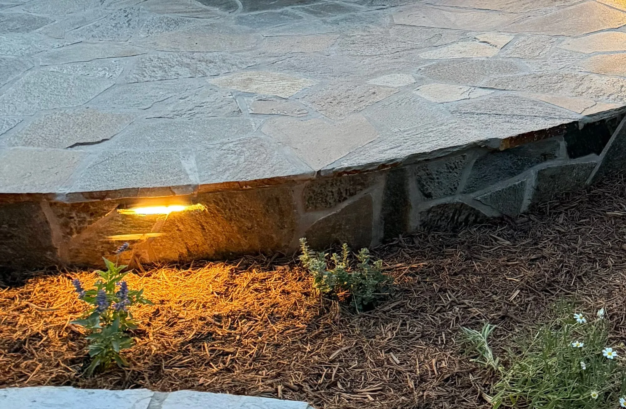 Edge of a raised flagstone patio with a light set into the stone face above a mulched planting bed